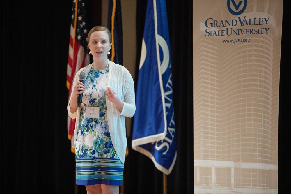Student presenting on stage, a GVSU banner and several flags behind her